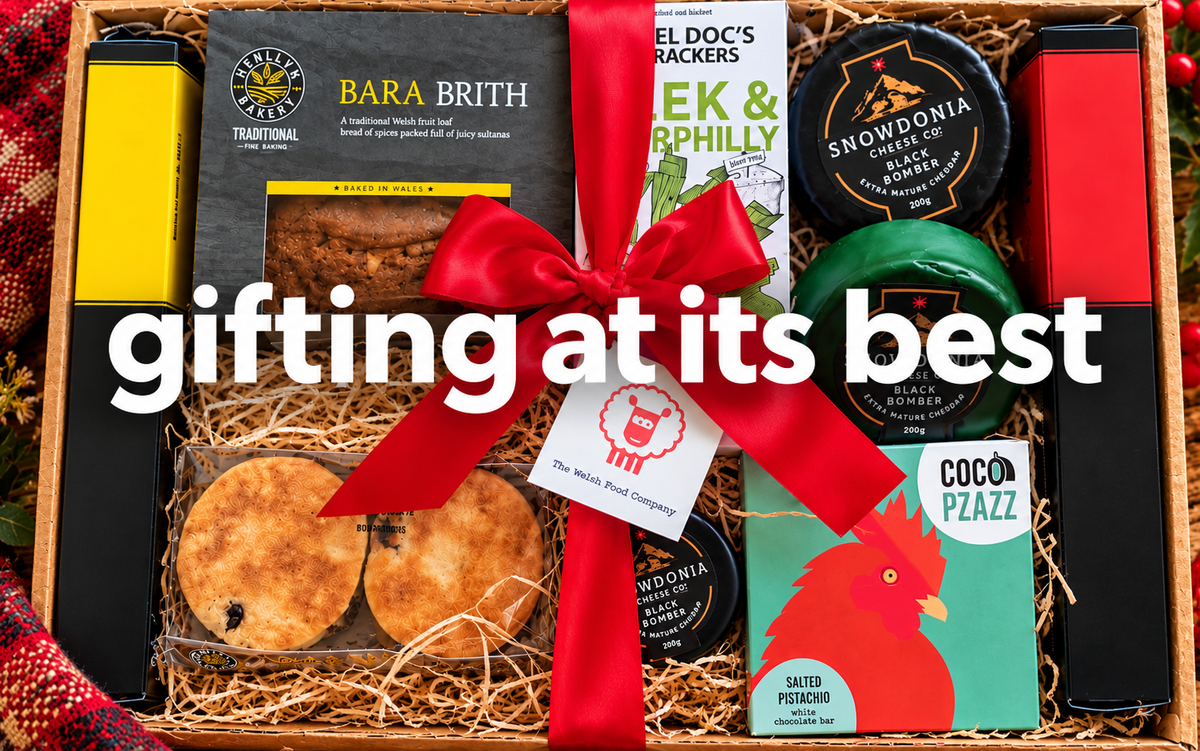 A Welsh Food gift box filled with assorted gourmet treats and packaged snacks, tied with a large red ribbon and overlaid with the text 'gifting at its best.' Visible items include a BARA BRITH cake, welsh cakes, two Snowdonia Cheese.